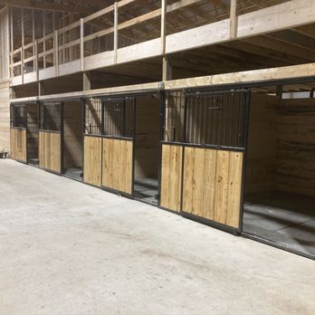 Blackstone Stables - Boarding Stalls - Inside a barn, several horse stalls are arranged in a row, each with wooden lower panels and metal upper bars. The stalls are part of a larger wooden structure with an upper level and open space above. The concrete floor is visible, and some tools and equipment are nearby, indicating ongoing construction or setup. - Horseback Riding Near Me - Howell, Michigan