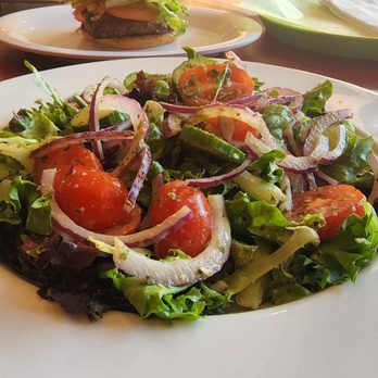 Project Burger - Salad - A fresh mixed salad with cherry tomatoes, sliced red onions, and assorted leafy greens, presented on a white plate. The salad appears to be lightly dressed and is set on a wooden table in a restaurant setting, with a burger and drink visible in the background. - American Near Me - Alameda, California