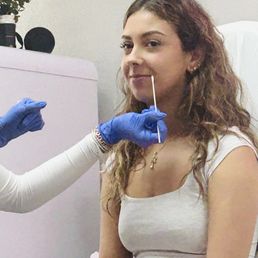 A woman with curly hair is seated while a healthcare worker in blue gloves administers a nasal swab test.