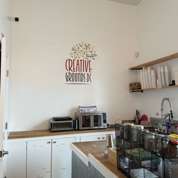 Creative Grounds DC - The image shows the interior of a coffee shop with a white wall featuring a colorful 'Creative Grounds DC' sign. There are wooden shelves holding disposable cups and a towel. The counter has a black wire organizer with supplies, a wooden block, and a coffee pitcher. Equipment such as a microwave, toaster, and small oven are on the counter. The space has a clean, minimalistic design with natural wood accents. - Art Classes Near Me - Washington, DC, District of Columbia