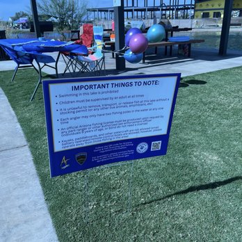 Painted Sky Park - Important notes - A blue informational sign at Painted Sky Park in Apache Junction, AZ, displays rules and regulations for lake activities, including prohibitions on swimming, requirements for supervision, and fishing license information. The sign is placed on a grassy area near a concrete walkway, with tables, balloons, and a water feature visible in the background. The setting appears to be a recreational outdoor space with amenities for visitors. - Parks Near Me - Apache Junction, Arizona