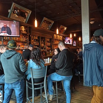 Sidecar - Full bar - The image depicts a bar interior with several patrons seated on high chairs around the bar counter. Multiple television screens are mounted on the wall, displaying different programs. The bar is stocked with various bottles of alcohol, and warm pendant lights hang from the ceiling, creating a cozy atmosphere. The ceiling has a textured pattern, and a concrete pillar is visible on the right side of the image. The patrons are engaged in conversations, and some are looking at the screens or their drinks. - Cocktail Bars Near Me - Denver, Colorado