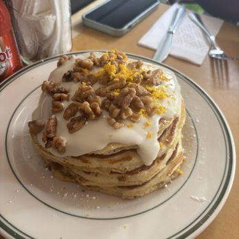 Sweet Chick - Lloyd's Carrot Cake Pancakes - A tall stack of fluffy pancakes is presented on a white plate with a green rim. The pancakes are topped with a generous layer of yogurt, sprinkled with chopped walnuts, and garnished with lemon zest. The setting includes a wooden table with utensils, a napkin, a smartphone, and a jar of sugar or sweetener in the background. - Southern Near Me - Brooklyn, New York