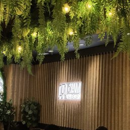Indoor space with a ceiling decorated with hanging green plants and string lights, and a wooden wall with a neon sign.