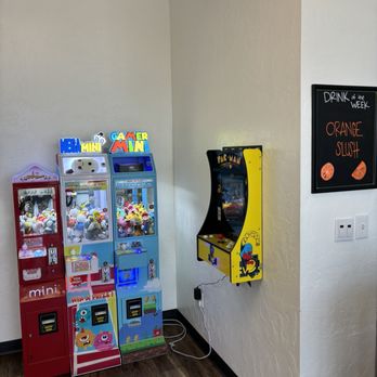 Boba Shack - Corner arcade! - The image shows three arcade claw machines, each filled with plush toys, positioned next to each other against a wall. The machines are colorful, with labels such as 'Mega Mini,' 'Gamer Mini,' and a red machine with a 'mini' label. To the right, a yellow Pac-Man arcade game is mounted on the wall, plugged into an outlet with visible cords. A black-framed sign on the wall advertises the 'Drink of the Week,' specifically an orange slush. The setting appears to be an indoor recreational area with a wooden floor and neutral-colored walls. - Bubble Tea Near Me - Apache Junction, Arizona