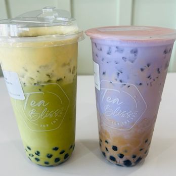 Tea Bliss - Two bubble tea drinks are placed on a white surface, each in a clear plastic cup with a lid. The left drink is green, likely matcha or green tea flavor, with tapioca pearls at the bottom. The right drink is purple with a pinkish hue, containing ice and tapioca pearls at the bottom. Both cups have a logo that reads 'Ten Bliss' and are set against a light-colored background. - Bubble Tea Near Me - Chandler, Arizona