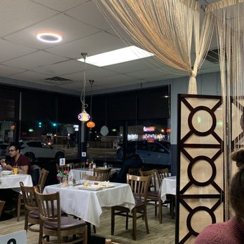 Bamyan Kabob - Inside dining - The image shows the interior of a restaurant during evening hours, with several tables covered in white tablecloths and wooden chairs arranged around them. There are small flower arrangements on some tables. The ceiling has hanging decorative curtains and colorful pendant lights. Large windows reveal a nighttime street scene outside with cars and illuminated signs. The setting appears casual and cozy, with a few patrons seated at tables. - Halal Near Me - Chicago, Illinois
