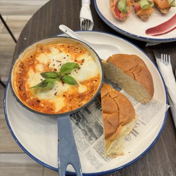 11:11 CAFE - Shakshuka - The image shows a meal served on a dark wooden table, including a small skillet of baked pasta with melted cheese and basil garnish, two slices of bread on a white plate, and a separate plate of appetizers topped with vegetables. The table setting includes a fork, knife, and napkin, with a background of a wooden floor. - Cafes Near Me - Brooklyn, New York