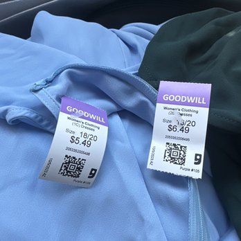 Goodwill - Same dress processed the same day, different prices. - Two women's dresses are shown with price tags at Goodwill. The light blue dress is labeled as size 18/20 and priced at $5.49, while the dark green dress is size 13/20 and priced at $6.49. The tags include QR codes and store information. The dresses are placed on a surface, possibly in a shopping cart or on a seat, with a dark background. - Thrift Stores Near Me - Phoenix, Arizona