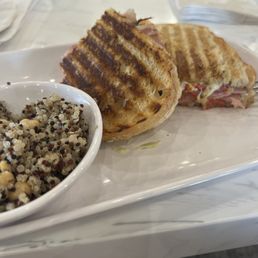 Rustic Italian panini and ancient grains - A grilled panini sandwich with ham and melted cheese is presented on a white plate, with visible grill marks on the bread. Next to it is a small bowl filled with a quinoa salad containing mixed grains and possibly chickpeas. The setting appears to be a casual dining environment, with a white table surface and napkins in the background.