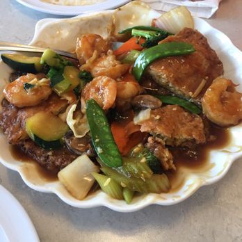 Dragon King - Shrimp egg foo young - A white, scalloped-edge plate contains shrimp egg foo young topped with mixed vegetables including zucchini, carrots, mushrooms, and snap peas, all covered in a savory sauce. Part of the dish is partially eaten, revealing the egg-based patty underneath. The plate is placed on a light-colored table, with a person's hand and a ring visible in the background, along with another partially eaten plate. - Chinese Near Me - Colorado Springs, Colorado