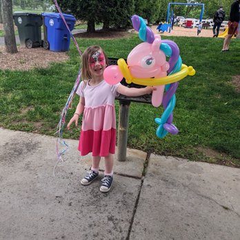 PartyyMagic - A girl with face paint and a pink dress stands next to a balloon unicorn sculpture at a park with playground equipment in the background. - Caricatures Near Me