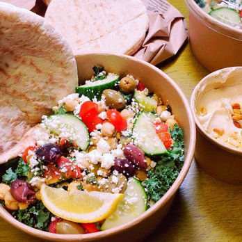 Suger Bōle - Mediterranean salad and hummus - A wooden table holds a large bowl of Mediterranean salad containing cucumbers, cherry tomatoes, black olives, feta cheese, and a lemon wedge. Next to the salad is a small cup of hummus topped with pine nuts and a drizzle of oil. A piece of pita bread is placed on the edge of the salad bowl, and another bowl of salad is partially visible in the background. The setting suggests a healthy, fresh meal with Middle Eastern influences. - Beer, Wine & Spirits Near Me - Biddeford, Maine
