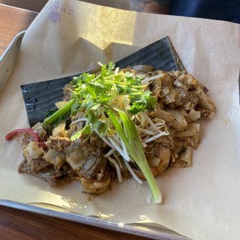 Charm Thai Eatery - A serving of stir-fried noodles with vegetables, including bean sprouts, green onions, and cilantro, is presented on a tray lined with parchment paper. The dish appears to include pieces of meat or tofu, and is garnished with fresh herbs. The tray is placed on a wooden table, with part of a person's arm and blue shirt visible in the background, indicating a casual dining setting at Charm Thai Eatery. - Thai Near Me - Westminster, Colorado