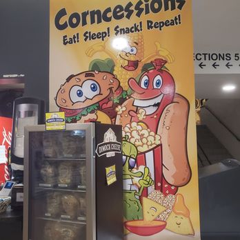 Corn Palace - A large, colorful poster displays cartoon food characters including a burger, hot dog, popcorn, and a corn cob, promoting concessions with the slogan 'Eat! Sleep! Snack! Repeat!'. The poster has a yellow background and is positioned behind a snack display case. The characters are animated and smiling, creating an inviting atmosphere for a snack stand or concession area. - Museums Near Me - Mitchell, South Dakota