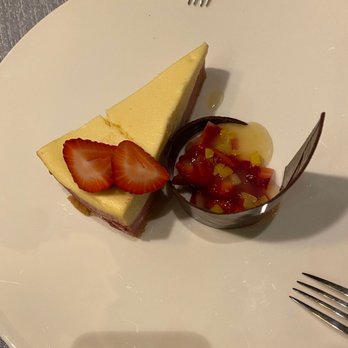 PY Steakhouse - Lemon raspberry cheesecake - A slice of lemon raspberry cheesecake garnished with strawberry slices and served with a fruit topping in a small cup on a white plate. - Steakhouses Near Me - Tucson, Arizona