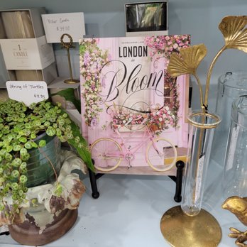 Self Indulgence - A table displays a variety of decorative items including a potted plant labeled 'String of Turtles,' a book titled 'London in Bloom' with pink floral cover art, a set of candles, and gold-colored ornaments. There are also glass containers and a small gold bird figurine, arranged in a retail or display setting. - Antiques Near Me - Troutdale, Oregon