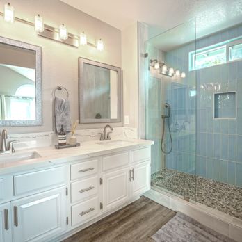 Youngren Construction - a bathroom with two sinks and a shower - The image shows a bathroom with a double vanity that has white cabinets and a marble countertop, two mirrors with decorative frames, and a row of four light fixtures above. To the right, there is a walk-in shower enclosed by a glass panel, with blue tiled walls, a built-in niche, and a pebble tile floor. A small horizontal window provides natural light. The flooring outside the shower appears to be wood or wood-like material, and a rug is placed in front of the vanity. - General Contractors Near Me - Fallbrook, California