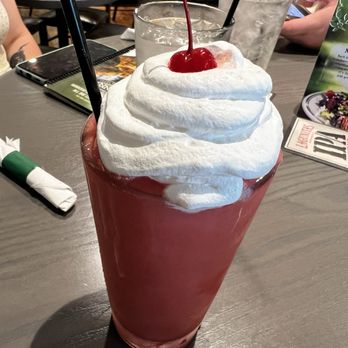 Bennigan's - A large strawberry milkshake with whipped cream and a cherry on top, served in a tall glass with two straws, on a restaurant table. - American Near Me - Urbandale, Iowa