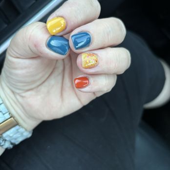 K Nails Spa - my nails - A hand with five painted nails is shown resting on a person's lap inside a vehicle. The nails are painted in different colors: yellow, blue, glittery gold, and orange. The person is wearing a white watch with a gold band on their wrist. The background includes part of the car interior and the person's black clothing. - Nail Salons Near Me - Plaistow, New Hampshire