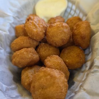 The Rock Restaurant - Corn nuggets - A basket lined with paper contains multiple fried corn nuggets, which are golden-brown and crispy. A small container of creamy dipping sauce is placed behind the nuggets. The presentation suggests a snack or appetizer, likely from a restaurant offering American or Mediterranean cuisine. - Greek Near Me - Normal, Illinois