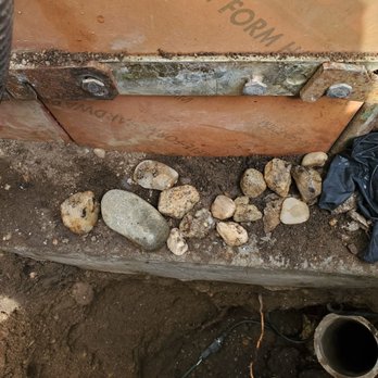 Prehoda Builders - More rocks - The photo shows a section of a construction or renovation site with various rocks and dirt on the ground. A metal component with bolts is attached to a wall or structure, and part of a pipe or conduit is visible at the bottom. Shoes are also visible in the lower part of the image, indicating a person standing at the site. - General Contractors Near Me