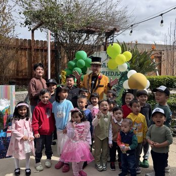 Top Class Magic - They loved the show! Amazing! - An outdoor children's birthday party features a magician performing for a group of kids. The magician is dressed in a yellow jacket and a top hat, holding a microphone, with balloons and a birthday banner behind him. The children are standing on a concrete patio, some smiling, laughing, and wearing colorful clothing. The setting includes a wooden fence, trees, and string lights overhead, indicating a backyard environment. - Magicians Near Me