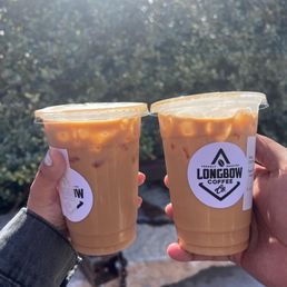 Butter beer latte (left) and pecan latte (right) - Two iced coffee beverages in clear cups with Longbow Coffee Co. labels are held outdoors in sunlight, with a blurred natural background.