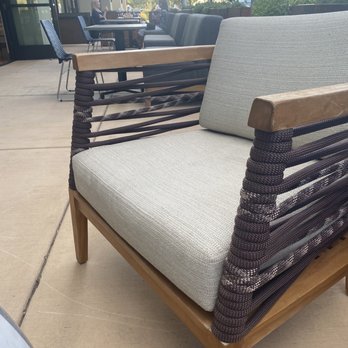 Crafted NM - Loved these chairs! Want them for myself - A contemporary outdoor chair features a wooden frame with woven dark brown sides and light-colored cushions. It is positioned on a concrete patio outside a restaurant, with additional tables and chairs, and patrons seated in the background. The setting includes glass windows, greenery, and trees, indicating a casual dining environment. - Wine Tasting Room Near Me - Albuquerque, New Mexico