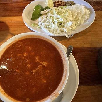 Jalapenos - Pozole 1/2 order with side garnish. This stew is amazing. - A bowl of red stew with visible chunks of meat is placed on a white plate with a spoon, alongside a plate of shredded cabbage, lime wedge, and spices. - Mexican Near Me - Schereville, Indiana