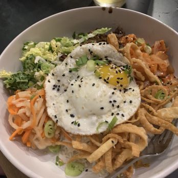 Block 15 Brewing - Kimchi Rice Bowl - A large white bowl contains a rice dish topped with a fried egg, black sesame seeds, and chopped green onions. The bowl also includes shredded vegetables, fried strips, and possibly kimchi. In the background, there is a glass of beer and a dark table surface, indicating a casual dining setting. - New American Near Me - Corvallis, Oregon
