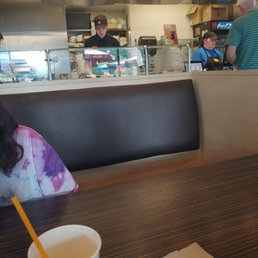 Awesome staff - The image shows a casual restaurant interior with customers at the counter. Staff members are visible behind a glass partition, preparing food and taking orders. The background includes a menu board and a refrigerator with beverages. The foreground features a table with a paper cup and a napkin, and part of a seated customer is visible. The setting appears to be a fast-casual dining establishment.
