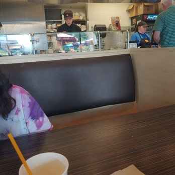 QDOBA Mexican Eats - Awesome staff - The image shows a casual restaurant interior with customers at the counter. Staff members are visible behind a glass partition, preparing food and taking orders. The background includes a menu board and a refrigerator with beverages. The foreground features a table with a paper cup and a napkin, and part of a seated customer is visible. The setting appears to be a fast-casual dining establishment. - Mexican Near Me - Duluth, Minnesota
