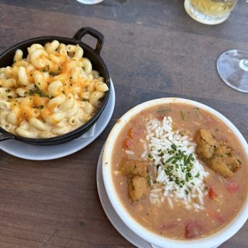 Subourbon - Gumbo and Mac and cheese - The image shows two bowls of food on a wooden table. One bowl contains macaroni and cheese topped with melted cheese and herbs, served in a small cast iron skillet. The other bowl contains gumbo with rice, vegetables, and fried chicken pieces, garnished with chopped green onions. In the background, there are drinks, including a glass of water, a glass of white wine, and a glass with a yellow beverage, along with a basket of green beans. The setting appears to be a casual dining environment, possibly a restaurant offering Southern-inspired cuisine. - Southern Near Me - Columbus, Ohio