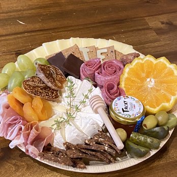Tricky Foods - Board made in class - A round cheese and fruit platter includes green grapes, orange slices, figs, chocolates, cured meats, and decorative herbs. The platter also has a honey dipper, a jar labeled 'Tucky', and various cheeses, arranged on a wooden table for serving. - Caterers Near Me - Madison, Wisconsin
