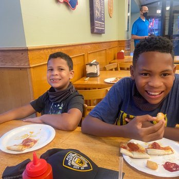 Gigio's Pizzeria - Pizza, Tater Tots - Two boys are sitting at a wooden table inside a casual pizzeria, smiling at the camera. The boy on the right is holding a piece of pizza, and both have plates with pizza slices and potato chips. The table has drinks with straws, and a police badge cap is also on the table. The background shows a staff member in a blue shirt and black mask, with restaurant decor and seating visible. - Pizza Near Me - Evanston, Illinois