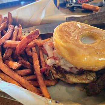 Jaspers Cafe - Glazed and confused - A burger topped with a glazed donut replacing the traditional bun, served with a side of French fries. The burger appears to contain cheese, bacon, and other fillings. The meal is presented on a metal tray lined with parchment paper, with a knife and some utensils visible in the background. - Burgers Near Me - Bluff Dale, Texas