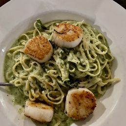 Scallop Pesto Linguine - A white round plate contains linguine pasta coated in green pesto sauce, garnished with chopped herbs. Four seared scallops with a golden-brown crust are placed on top of the pasta, arranged with some scallops near the edges and others in the center. The dish appears to be served in a restaurant setting, emphasizing seafood and pasta cuisine.