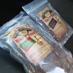Best beef jerky ever in the world hands down the only place I will get beef jerky from for the rest of my life!!!! - Two sealed packages of Mike's Jerky, a beef jerky product, with Christmas-themed labels, placed on a car seat.