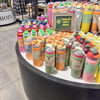 Public Lands - A retail store display showcases numerous reusable water bottles in various pastel and bright colors, arranged on a round table. The bottles have flip-top lids and are grouped by color. In the background, clothing racks with garments and mannequins are visible, indicating a clothing and accessories section within the store. A sign on the table reads 'GREAT GIFT IDEA,' suggesting these bottles are marketed as gift options. - Sports Wear Near Me - Framingham, Massachusetts