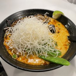 Red Curry Ramen - A bowl of red curry ramen with crispy noodles, green vegetables, and a lime wedge, served in a black bowl on a white table.