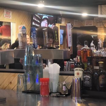 Roosters - Beer - The image depicts a bar counter with various bottles of alcohol, including Jameson and Jack Daniel's, and glassware. A bartender is visible behind the counter, preparing drinks or serving customers. Several glasses are stacked upside down on the counter, and a container with straws and a red condiment jar are also present. In the background, patrons are seated, and a television screen is mounted on the wall, showing a person’s face. The setting appears to be a lively bar or pub environment. - Chicken Wings Near Me - Maumee, Ohio