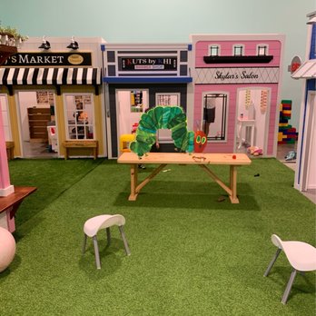 Wisco Kidz Place - The image shows a children's play area with artificial grass flooring, small white chairs, and a wooden table. Behind the setup are miniature storefront facades with signs, including a barber shop, a salon, and a market, decorated with colorful elements and props. The scene is designed for imaginative play and is part of an indoor space for children. - Playgrounds Near Me - Madison, Wisconsin