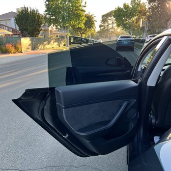 Rey's Window Tinting - The photo captures the open rear passenger door of a modern vehicle parked on a street. The interior door panel features a combination of leather and fabric materials, with a window partially rolled down. The background shows a residential neighborhood with trees, houses, and other parked cars, illuminated by sunlight, indicating early morning or late afternoon. - Auto Glass Services Near Me - Brea, California