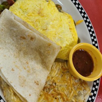 Ze's Diner - Fiesta omelette and substituted the tortilla and set up the bread - A breakfast dish served on a white plate with a black and white checkered border, including a yellow omelette, a folded flour tortilla, crispy hash browns, and a small yellow cup of salsa. The plate is placed on a red table surface, typical of a 1950s-inspired diner setting. - Diners Near Me - Eagan, Minnesota