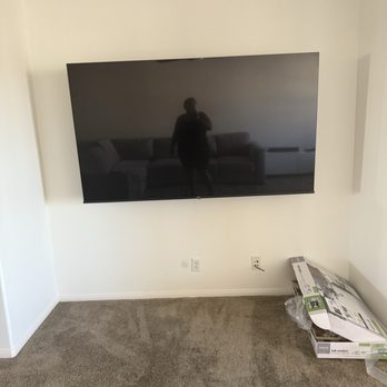 99 TV Install - Here, he mounted a 75" tv and installed an outlet behind it - A flat-screen TV is mounted on a white wall in a room with beige carpet. There are electrical outlets below the TV and boxes of TV mounting hardware on the floor. - Home Theatre Installation Near Me - Upland, California