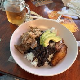 Lechon Bowl - A bowl containing rice, black beans, shredded pork, sliced avocado, and fried plantain, with a beer in a mason jar on a wooden table.