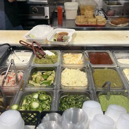 A self-serve condiment station at a Mexican restaurant features guacamole, shredded cheese, salsas, and fresh vegetables like jalapeños and lime wedges.