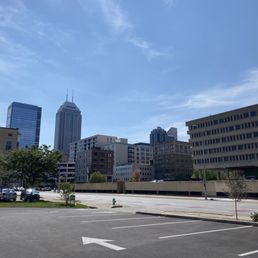 Rare downtown parking lot for Harrison's - The image shows a city skyline with multiple tall buildings, including a prominent skyscraper with antennas, under a bright, mostly clear blue sky. In the foreground, there is an empty parking lot with marked parking spaces, a few small trees, and a fire hydrant. The scene suggests a quiet urban environment during daytime.