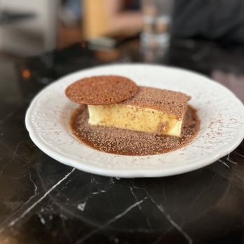 Geppetto - Tiramisu - A slice of tiramisu dessert featuring layers of mascarpone cheese and coffee-soaked sponge cake, dusted with cocoa powder, is presented on a white ceramic plate. A round chocolate cookie rests on top of the tiramisu, and the plate is set on a dark marble table. The background shows a blurred indoor setting with a glass of water and other objects. - Italian Near Me - Cambridge, Massachusetts
